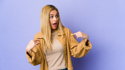 Young blonde woman isolated on purple background surprised pointing with finger, smiling broadly.