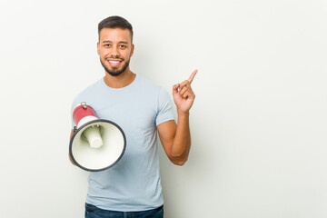 Young mixed race asian man holding a megaphone smiling cheerfully pointing with forefinger away.