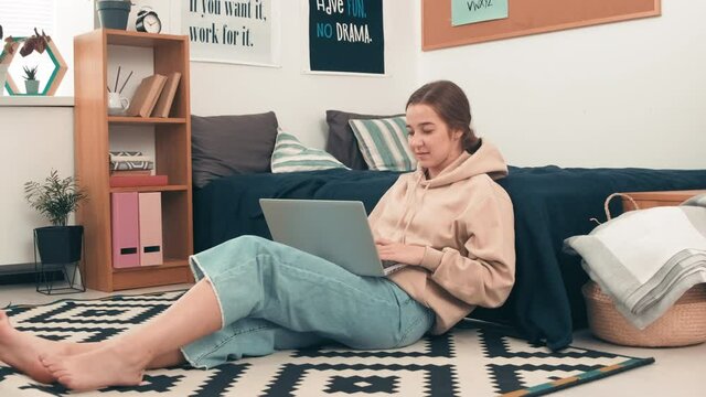 Handheld dolly-in shot of cheerful girl sitting on floor in cozy college dormitory and typing on laptop while doing her assignment