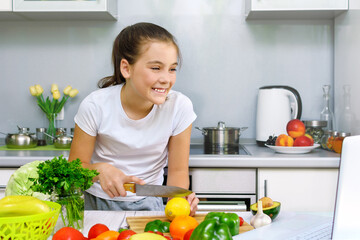 Happy beautiful young girl enjoying cooking looking for recipe on laptop in kitchen. Food blogger concept