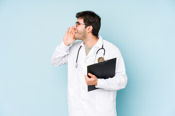 Young doctor man isolated on blue background shouting and holding palm near opened mouth.