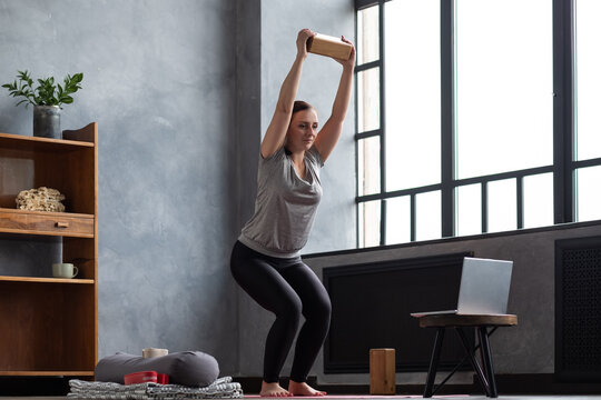 Caucasian woman doing Utkatasana, Chair Pose using block at home during online lesson.