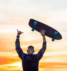 Silhouette of young sporty man enjoying carefree life on sunset or sunrise. Holds surf skateboard in right hand and showing shaka sign with left hand. Summertime chill. Outdoors sport activity © Creative Cat Studio