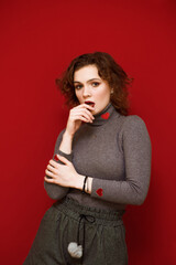 Portrait of an attractive girl with curly hair on a red background, looks into the camera with a serious face and poses, wearing gray clothes. Pretty lady with focused face posing on red background.