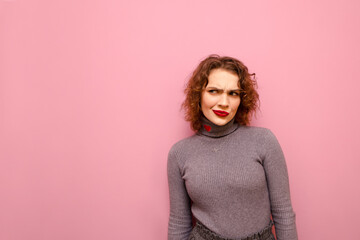 Portrait of suspicious lady in gray sweater and with curly red hair on pink background, looks away at copy space with pensive face. Suspicious pretty girls isolated on pink background.