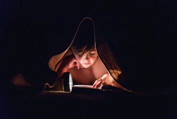 Young boy reading a book under a blanket using a flashlight.