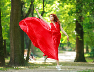 Young fashionable woman in long red luxurious dress walks in summer park