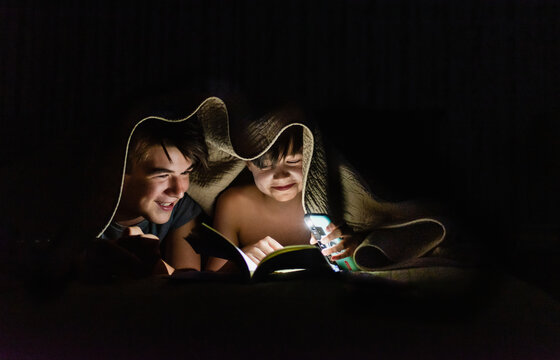 Brothers reading a book under a blanket by flashlight at night.