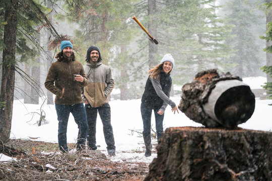 Young woman throws axe at a target in the snowy woods while her friends watch