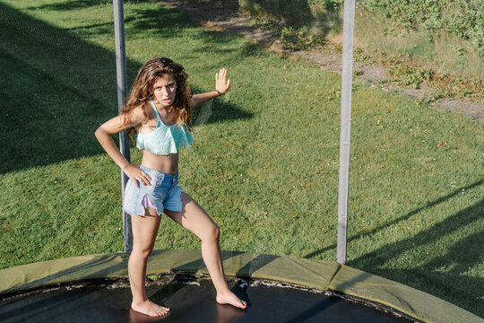 Horizontal photo of young brunette teenager on a trampoline leaning with her hand on the net on a park. The girl wears short jeans and a top and looks tired, passive and without energy.