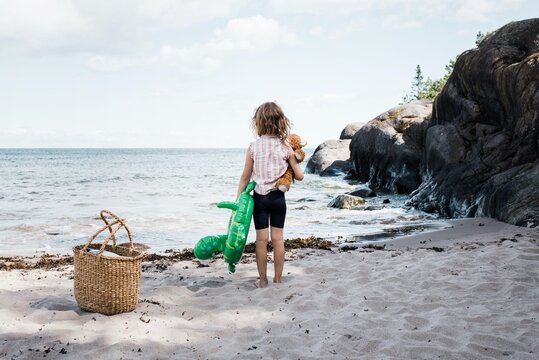 girl stood on the beach holding her bear and inflatable by the sea