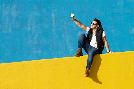 Young bearded man with sunglasses sitting on yellow wall,taking selfie