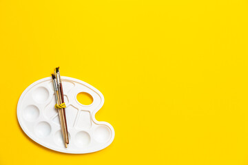 Clean plastic white palette with wooden natural paint brushes on yellow background. Banner with place for text for art school, hobbies, drawing lessons, expressing creativity or lack of inspiration