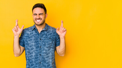 Young caucasian man isolated on yellow bakground crossing fingers for having luck