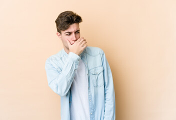 Young caucasian man isolated on beige background thoughtful looking to a copy space covering mouth with hand.