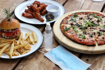 
New normal in restaurants, view of fast food pizza, hamburger and chicken wings with protection mask and antibacterial gel