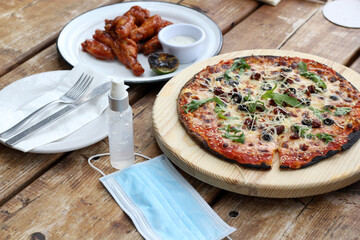 
New normal in restaurants, view of fast food pizza, hamburger and chicken wings with protection mask and antibacterial gel