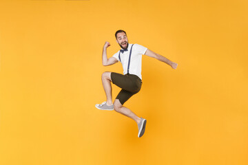 Full length side view portrait of excited young bearded man 20s wearing white shirt suspender shorts posing jumping clenching fists doing winner gesture isolated on bright yellow background studio.