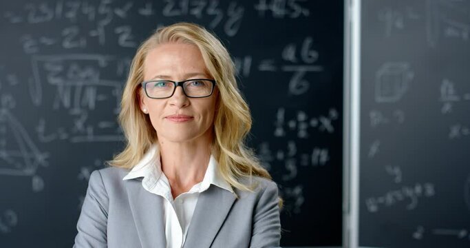 Portrait of smiled happy beautiful woman teacher in glasses. Blackboard with formulas, math and physic laws. Mathematics lesson. Caucasian female lecturer smiling to camera in school. Teaching concept