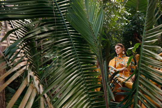 Stylish woman stands among large tropical palm plants.