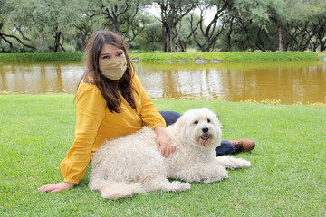 
latin woman with protection mask sitting on the grass with white dog, walking on the shore of the lake, new normal