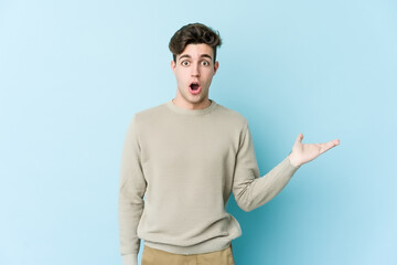 Young caucasian man isolated on blue background impressed holding copy space on palm.