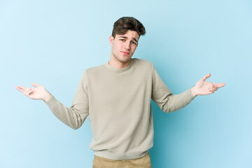Young caucasian man isolated on blue background doubting and shrugging shoulders in questioning...