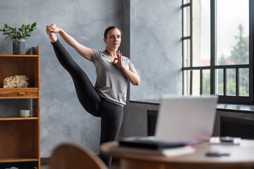Young flexible woman balancing doing asana Standing Hand to Toe at home during online lesson.