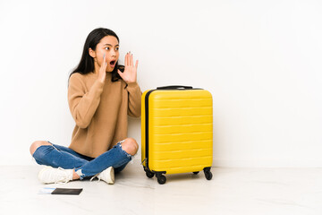 Young chinese traveler woman sittting on the floor with a suitcase isolated shouts loud, keeps eyes opened and hands tense.