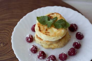 Juicy curd cheesecakes lie on a white saucer with raspberries and mint leaf. Light background