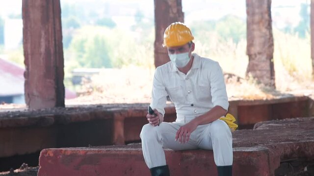 Resting place in the shade at the construction site. A male worker in a mask sits down to relax physical strength during construction work.