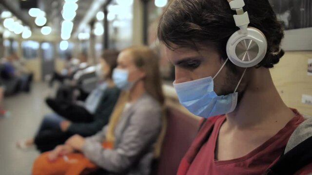 Side view of young brunette male wearing protective mask and listening to music in headphones while sitting in underground subway during covid-19 outbreak, lockdown, public caution, social distancing