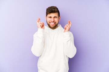 Young caucasian man isolated on purple background crossing fingers for having luck