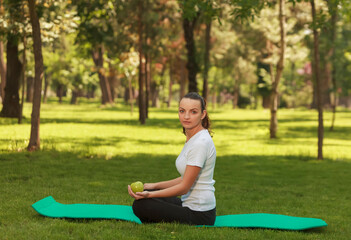 Fototapeta premium Young woman with a green apple in her left hand is relaxing in a morning on a mat outdoors in a park