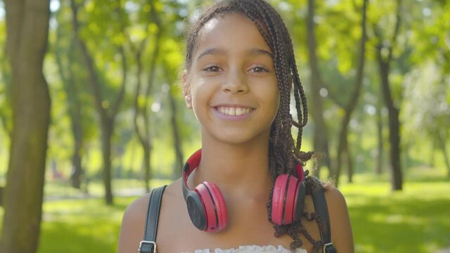 Close-up portrait of charming happy girl with afro pigtails and brown eyes taking off headphones and smiling at camera. Cheerful African American child posing in sunlight in summer park.