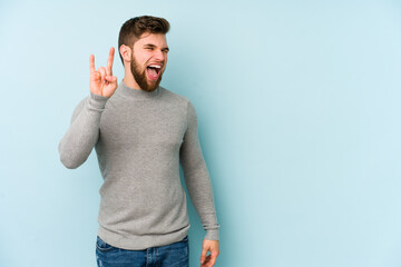 Young caucasian man isolated on blue background showing a horns gesture as a revolution concept.