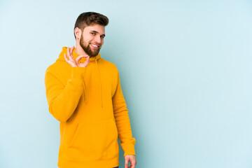 Young caucasian man isolated on blue background winks an eye and holds an okay gesture with hand.