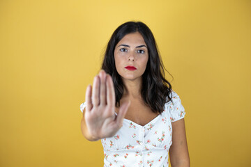 Beautiful woman over yellow background serious and doing stop sing with palm of the hand.