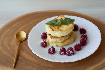 Juicy curd cheesecakes lie on a white saucer with raspberries and mint leaf. With golden spoon. Light background