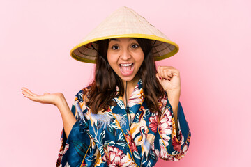 Young mixed race hispanic woman isolated holds copy space on a palm, keep hand over cheek. Amazed and delighted.