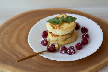 Juicy curd cheesecakes lie on a white saucer with raspberries and mint leaf. Light background