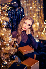 Young beautiful woman in elegant evening dress sitting on floor near christmas tree and presents on a new year eve. Interior with christmas decorations. Shallow DOF and bokeh background.