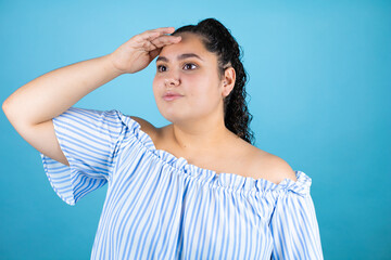 Young beautiful woman with curly hair over isolated blue background very happy and smiling looking far away with hand over head. Searching concept.