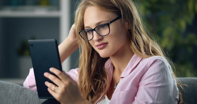Attractive lovely girl with blond hair using digital tablet while sitting on cozy sofa. Happy woman in casual clothing and eyewear spending free time online.