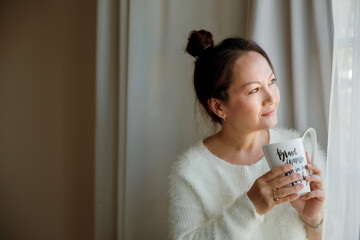 A happy and content woman reflecting next to the window at home