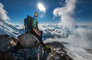 Climber in a safety harness, helmet, and high mountaineer boots with picturesque clouds background sitting at 3600m altitude on big rock and looking at summit during Mont Blanc ascending, France route - obrazy, fototapety, plakaty