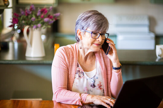 Mature woman on laptop at home using mobile phone to call for tech support