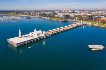 Aerial view of a long straight jetty with buildings on the end and a coastal city in the background