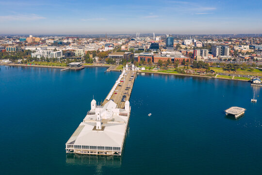 Aerial view of a long straight jetty with buildings on the end and a coastal city in the background