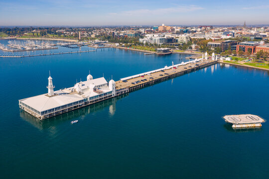 Aerial view of a long straight jetty with buildings on the end and a coastal city in the background